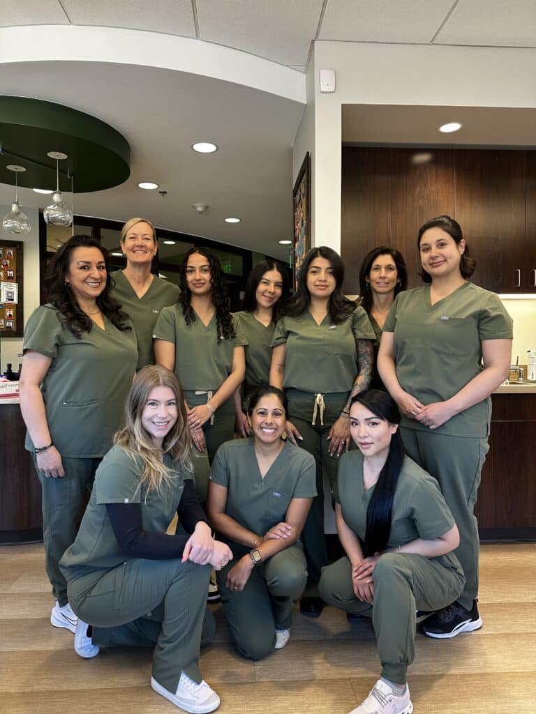 Kulkarni Orthodontics team smiling together inside the office, wearing matching olive-green scrubs in a bright, welcoming clinical space