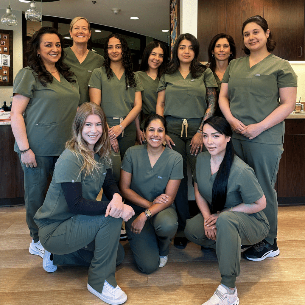 Kulkarni Orthodontics team smiling together inside the office, wearing matching olive-green scrubs in a bright, welcoming clinical space